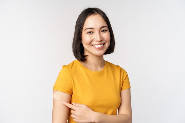 Covid19 vaccination campaign Smiling asian girl pointing at her band aid on shoulder recommending get vaccine from omicron coronavirus white background