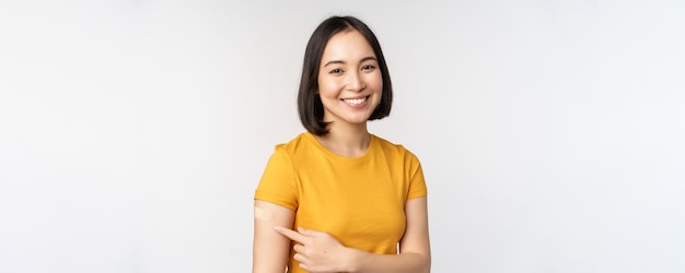 Free photo covid19 vaccination campaign smiling asian girl pointing at her band aid on shoulder recommending get vaccine from omicron coronavirus white background