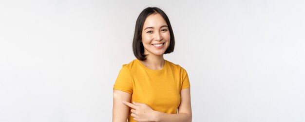 Covid19 vaccination campaign Smiling asian girl pointing at her band aid on shoulder recommending get vaccine from omicron coronavirus white background