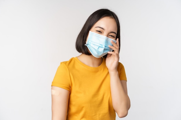 Covid19 예방 접종 및 의료 개념 배경 위에 서 있는 코로나바이러스 백신 접종 후 반창고를 보여주는 의료용 얼굴 마스크를 쓴 귀여운 아시아 소녀