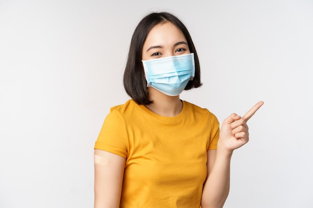 Covid19 예방 접종 및 의료 개념 의료 마스크를 쓴 귀여운 아시아 소녀의 초상화는 코로나바이러스 백신이 배경 위에 서 있는 후 어깨에 반창고를 하고 있습니다