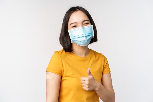 Covid19ワクチン接種とヘルスケアの概念医療マスクのかわいいアジアの女の子の肖像画は、白い背景の上にコロナウイルスワクチンが立った後、肩にバンドエイドを持っています