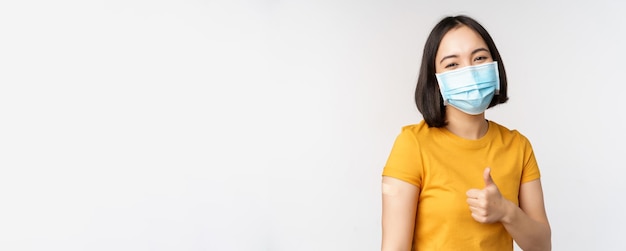 Covid19 vaccination and healthcare concept Portrait of cute asian girl in medical mask has band aid on shoulder after coronavirus vaccine standing over white background