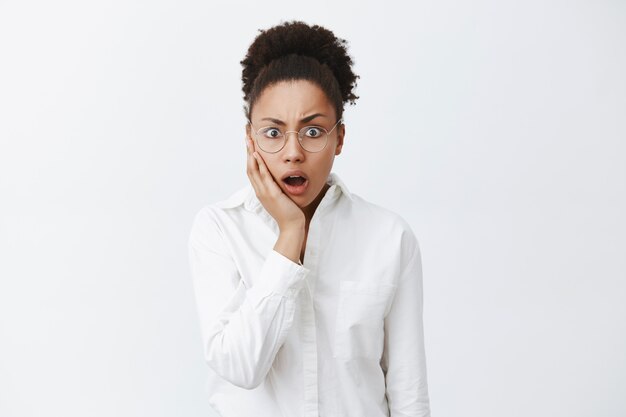 Coworker came work in blood, woman being stunned and shook. Portrait of shocked intense african-american female employer in white-collar shirt dropping jaw and gasping, staring amazed over grey wall