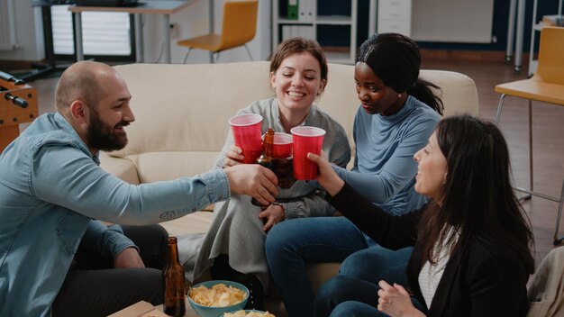 Коллеги звенят чашками и бутылками, наслаждаясь отдыхом после работы. Счастливые коллеги подбадривают с алкогольным напитком, чтобы отпраздновать вечеринку, веселиться и развлекаться в нерабочее время в офисе.