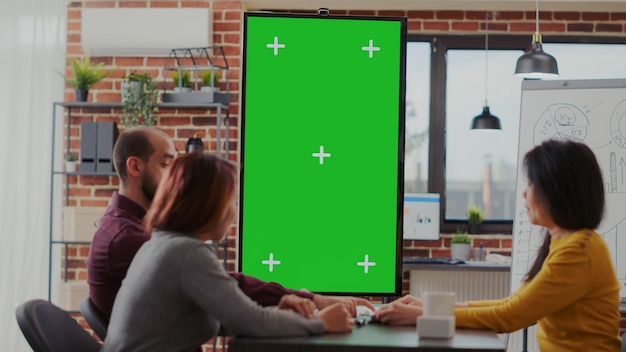 비즈니스 전략을 계획하기 위해 수직으로 모니터에 녹색 화면이 있는 사무실에서 회의를 하는 동료. 격리된 템플릿과 크로마 키가 있는 모형 복사 공간을 보고 있는 다양한 사람들의 팀.
