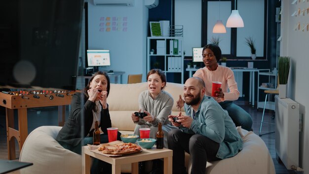 Coworkers playing video games with controllers on console after work. Cheerful colleagues enjoying game play with joysticks on television, having fun with beer bottles and snacks.