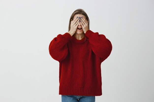 Coworkers preparing surprise for birthday girl. portrait of beautiful mature caucasian woman in red loose sweater, covering eyes with palms and opening mouth, waiting for gist or playing hide and seek