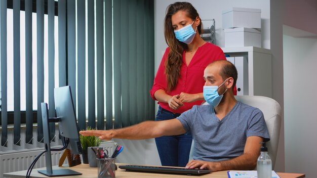 Сотрудники в защитных масках работают вместе на рабочем месте во время пандемии. Команда в новом обычном офисном рабочем пространстве в личной корпоративной компании печатает на клавиатуре компьютера, глядя на рабочий стол