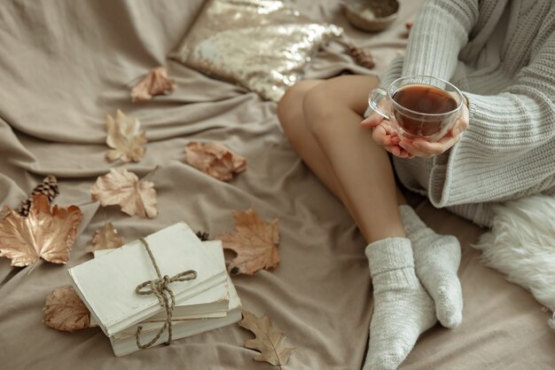 Cozy autumn background with female legs in warm socks, a cup of tea and autumn leaves in bed.