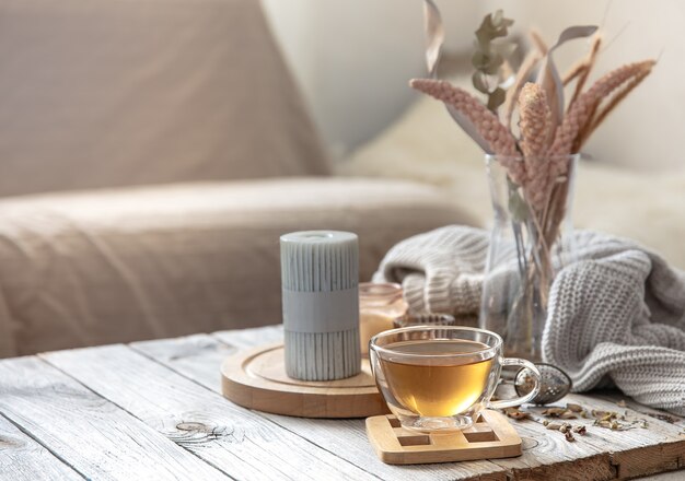 Уютная осенняя домашняя композиция с чашкой чая, свечами и вязанным элементом на размытом фоне интерьера комнаты.