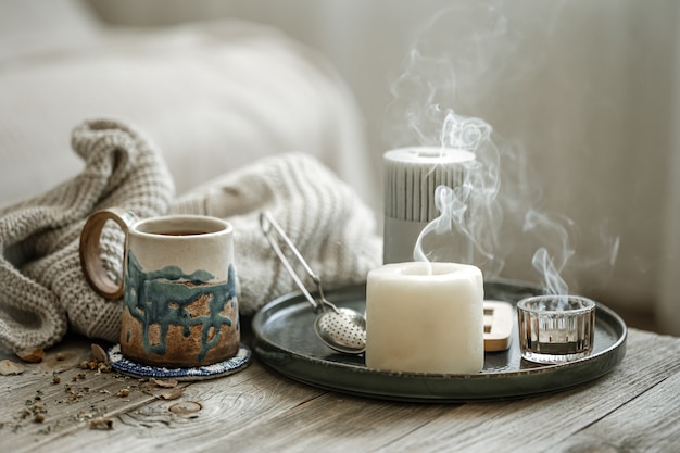 Foto gratuita composizione accogliente con una tazza in ceramica, candele e un elemento a maglia su uno sfondo sfocato.