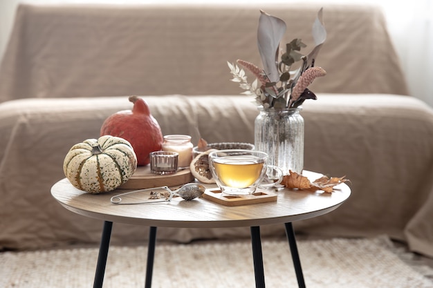 Accogliente natura morta domestica con una tazza di tè, zucche, candele e dettagli decorativi autunnali su un tavolo su uno sfondo sfocato della stanza.
