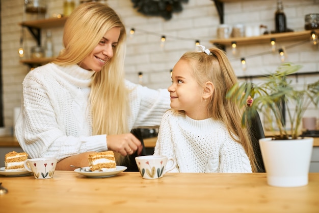 Cozy image of happy young mother with long blonde hair posing in kitchen with her adorable daughter, sitting at table, having tea and eating cake, looking at each other and smiling, talking