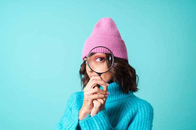 無料Photo ニットの青いセーターとピンクの帽子をかぶった若い女性の居心地の良い肖像画は、拡大鏡を持って、浮気し、楽しんで明るい化粧をしています