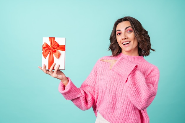 Free photo cozy portrait of a young woman in a pink knitted sweater and with bright makeup on turquoise with a gift box with a red bow