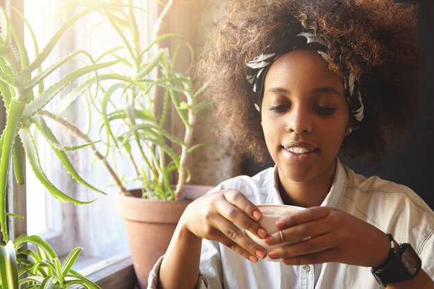 Cozy shot of African hipster girl wearing bandana and ring in her nose, holding cup of coffee or tea, having hot drink on winter morning while sitting alone at nice cafeteria or in her kitchen at home