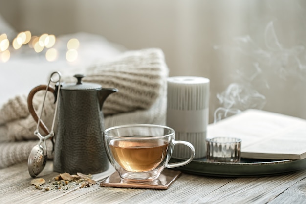 Cozy still life with a glass cup of tea, a teapot and candles on a blurred background with bokeh.