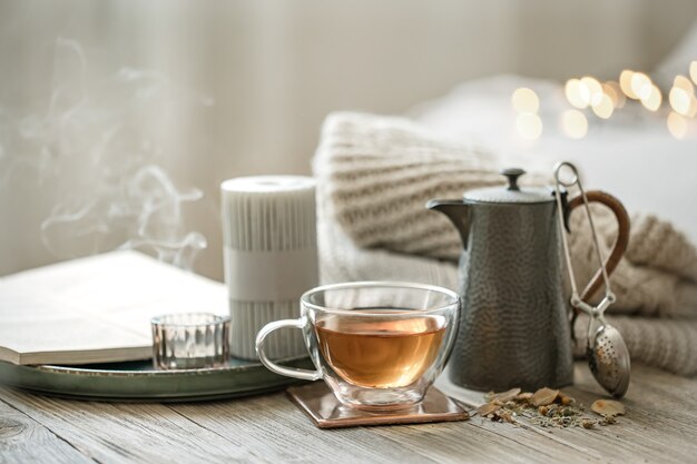 Cozy still life with a glass cup of tea, a teapot and candles on a blurred background with bokeh.