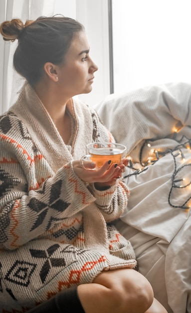 Foto gratuita accogliente tazza di tè trasparente a una ragazza carina in un caldo maglione lavorato a maglia contro la finestra