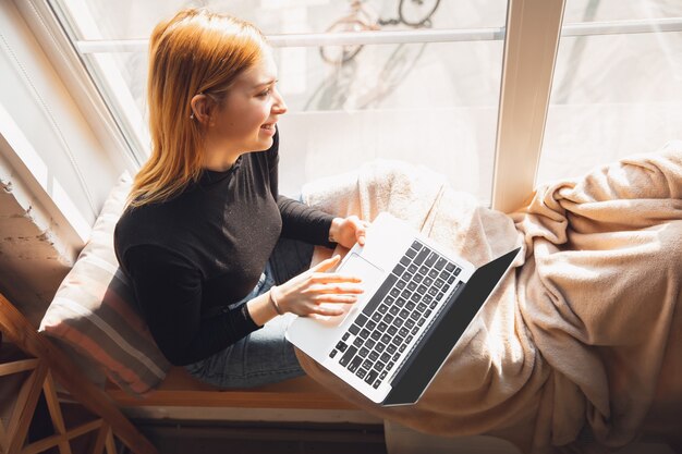 Cozy and warm. Young woman using gadgets to watch cinema, photos, online courses, taking selfie or vlog, online shopping or surfing. Reading news, has online conference, education, meeting friends.