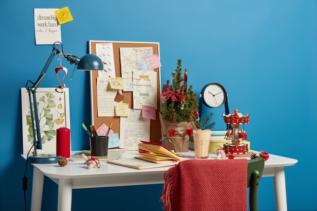 Cozy work space with decorated Christmas tree, traditional winter drink, board with sticky notes to remember, desk lamp for good lightening.