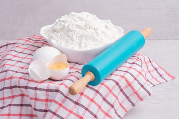 Cracked egg, rolling pin and bowl of flour on tablecloth.