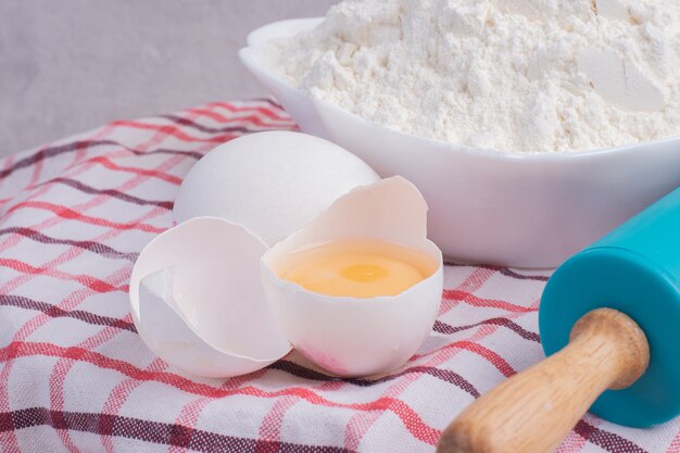 Cracked egg, rolling pin and bowl of flour on tablecloth.