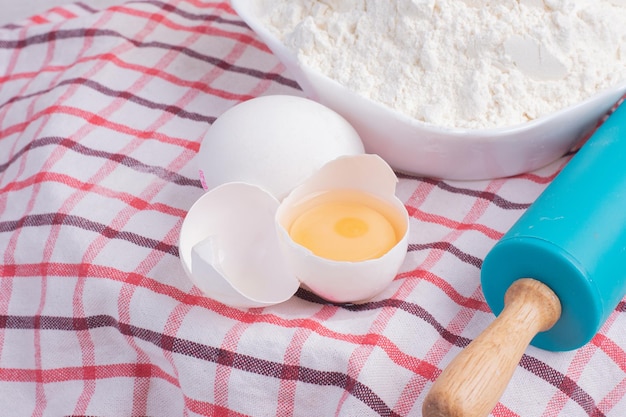 Cracked egg, rolling pin and bowl of flour on tablecloth.