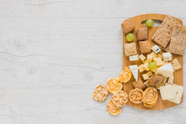 Cracker, cheese blocks, grapes and crisp bread and cookies on chopping board over the desk