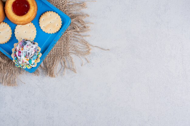 Crackers, cupcakes and jelly-filled cakes on a blue platter on marble table.