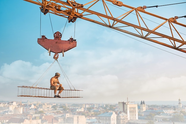Crane holding iron construction, where sitting builder with bare torso, eating and drinking milk. Extreme building on high. Cityscape on background.
