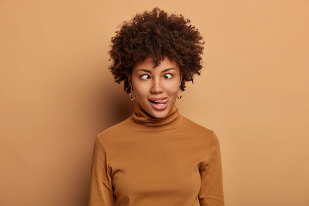 Foto gratuita la pazza donna afroamericana fa una faccia buffa, tira fuori la lingua e incrocia gli occhi, scherza, indossa un dolcevita marrone casual