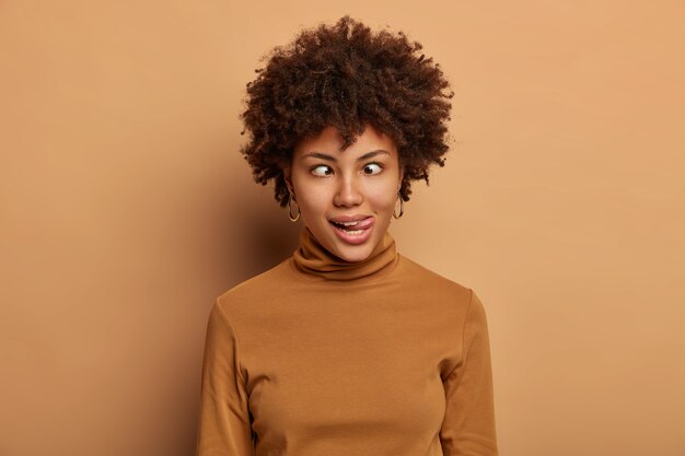 Crazy Afro American woman makes funny face, sticks out tongue and crosses eyes, fools around, wears casual brown turtleneck