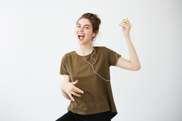 クレイジー陽気な若い女性が白い背景の上にヘッドフォンで音楽を聴いて歌って踊る。