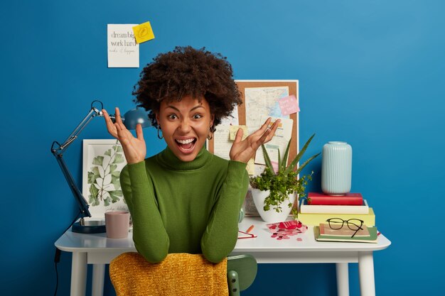 Crazy dark skinned young woman raises hands with annoyance, wears casual clothes, feels tired, has deadline, poses on chair near white table, isolated over blue background.