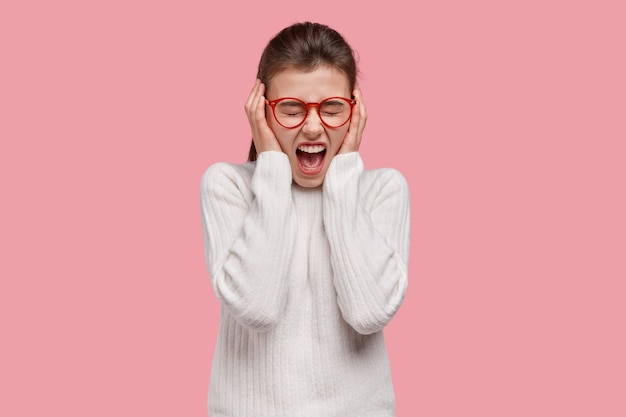 Free photo crazy desperate woman covers ears, shouts angrily, being emotional, annoyed with loud noise, wears white casual outfit