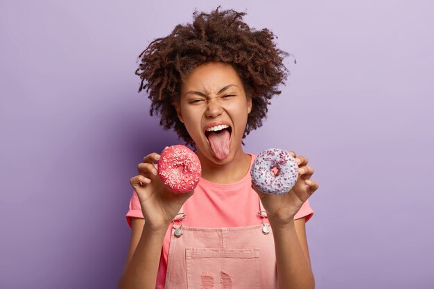 Crazy emotional dark skinned teenage girl shows tongue, holds two tasty doughnuts