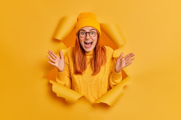 Crazy emotional redhead young woman raises palms and exclaims loudly thrilled by hearing great news wears yellow hat knitted jumper.