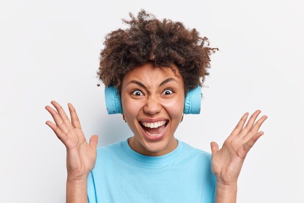 クレイジーな感情的な女性は手のひらを上げたまま叫び声は大声で反応しますカジュアルな青いTシャツに身を包んだ素晴らしいニュースに反応します白い背景の上に分離されたヘッドフォンを介して音楽を聴きます人間の反応の概念