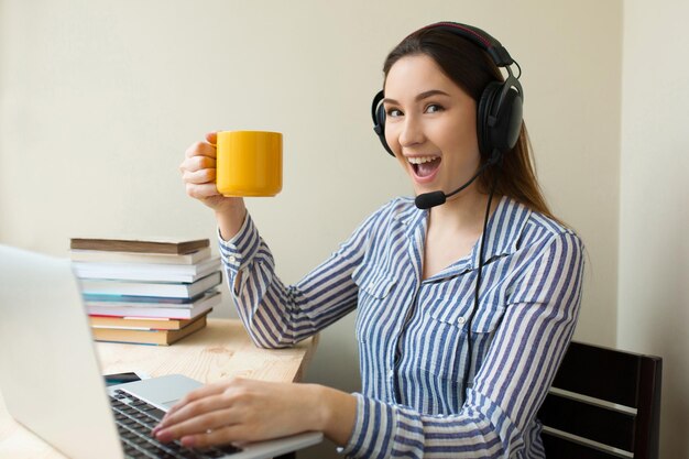 Crazy freelance operator working online with headsets and a laptop in a desktop at office. Happy call centre woman working from home talking with customer and drinking coffee