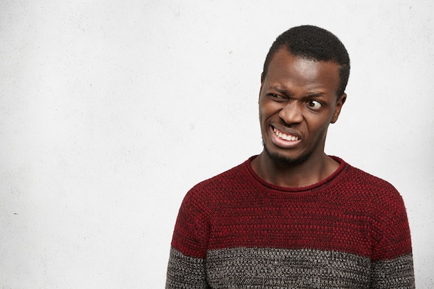 Pazzo divertente giovane afroamericano che indossa maglione casual in posa al chiuso facendo smorfie, facendo bocca, stringendo i denti e ammiccanti. espressioni facciali umane, emozioni e sentimenti