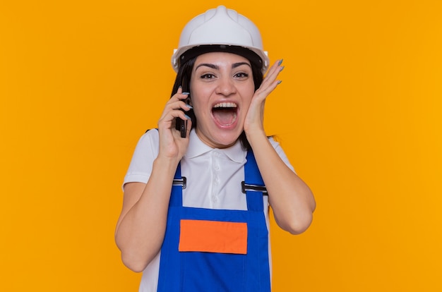 오렌지 벽 위에 서있는 휴대 전화에 얘기하는 건설 유니폼과 안전 헬멧에 미친 행복하고 흥분된 젊은 작성기 여자