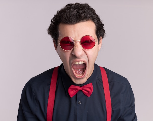 Crazy mad stylish man with bow tie wearing glasses and suspenders shouting with aggressive expression standing over white wall