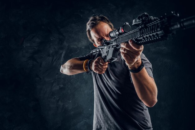 Crazy middle-aged man dressed in casual clothes holding an assault rifle and aims at the target. Studio photo against a dark textured wall