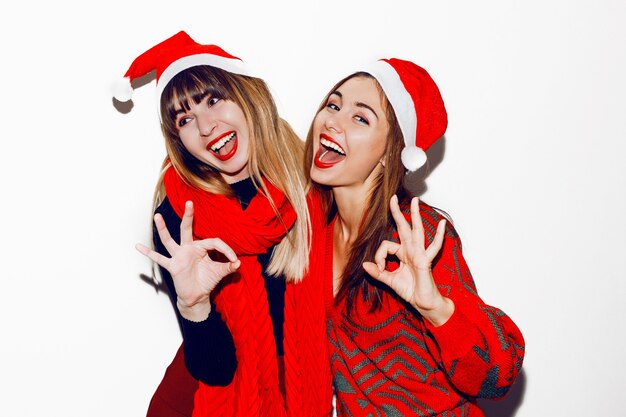 Crazy new year party mood. Two drunk laughing women having fun and posing in cute masquerade hats. Red sweater and scarf. Showing ok by hands.