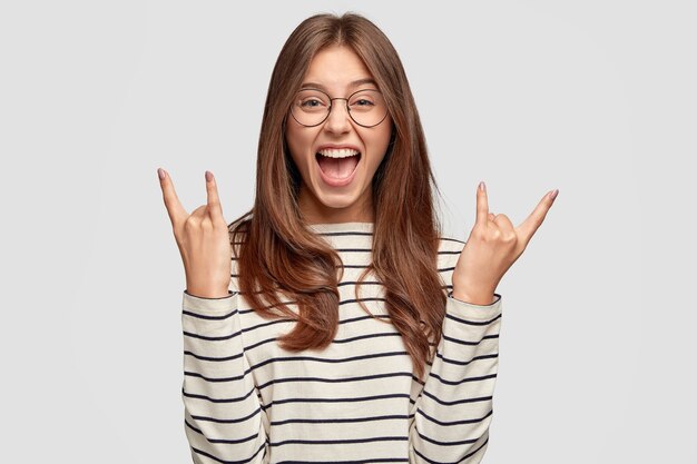 Crazy overjoyed woman makes rock n roll gesture, wears transparent glasses, striped sweater, models against white wall. Smiling female rocker gestures indoor alone. Horn gesture concept