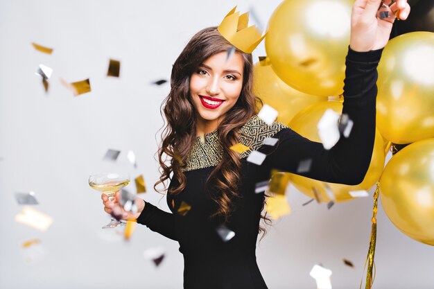 Crazy party time of beautiful woman in elegant black dress and yellow crown celebrating new year, birthday, having fun, dancing, drinking alcohol cocktails.Emotion face, red lips, gold balloons.