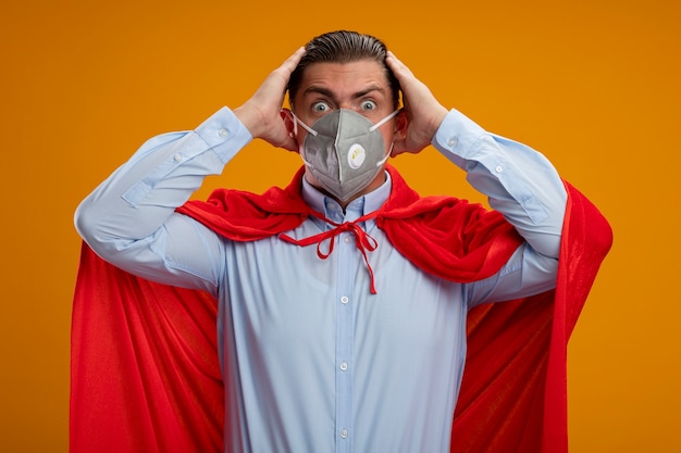 Crazy super hero businessman in protective facial mask and red cape looking at camera with crazy amazed look of surprise holding hands on his head standing over orange background