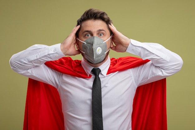 Crazy super hero businessman in protective facial mask and red cape  with crazy amazed look of surprise holding hands on his head standing over green wall